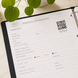 My Medical Folder open to the Personal Information section, showing personal details, emergency contact information and a photo template QR code on a marble surface with green leaves.