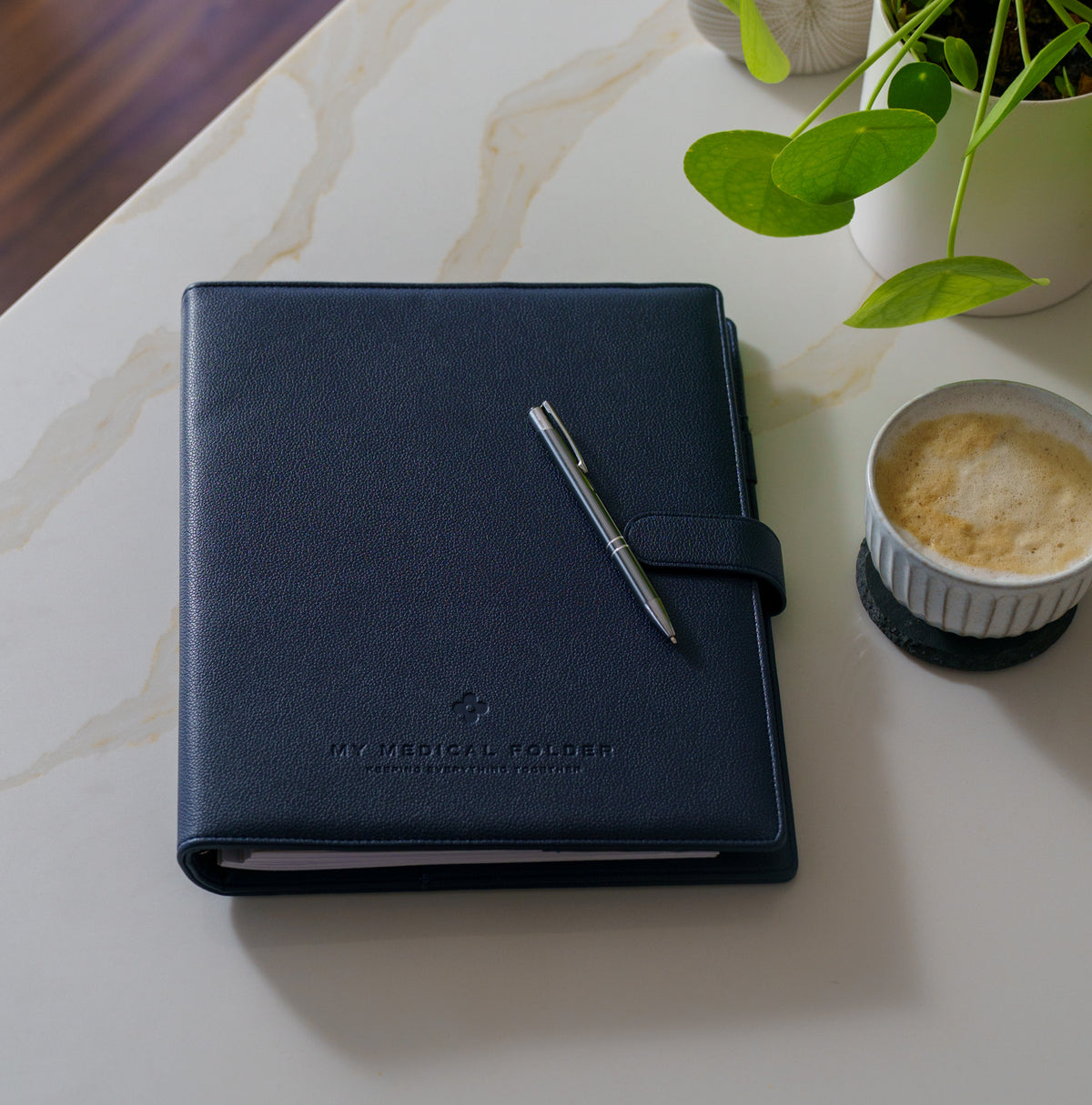 Navy My Medical Folder medical organiser on a marble tabletop beside a coffee and plant, designed to organise medical records, appointments and important health documents.