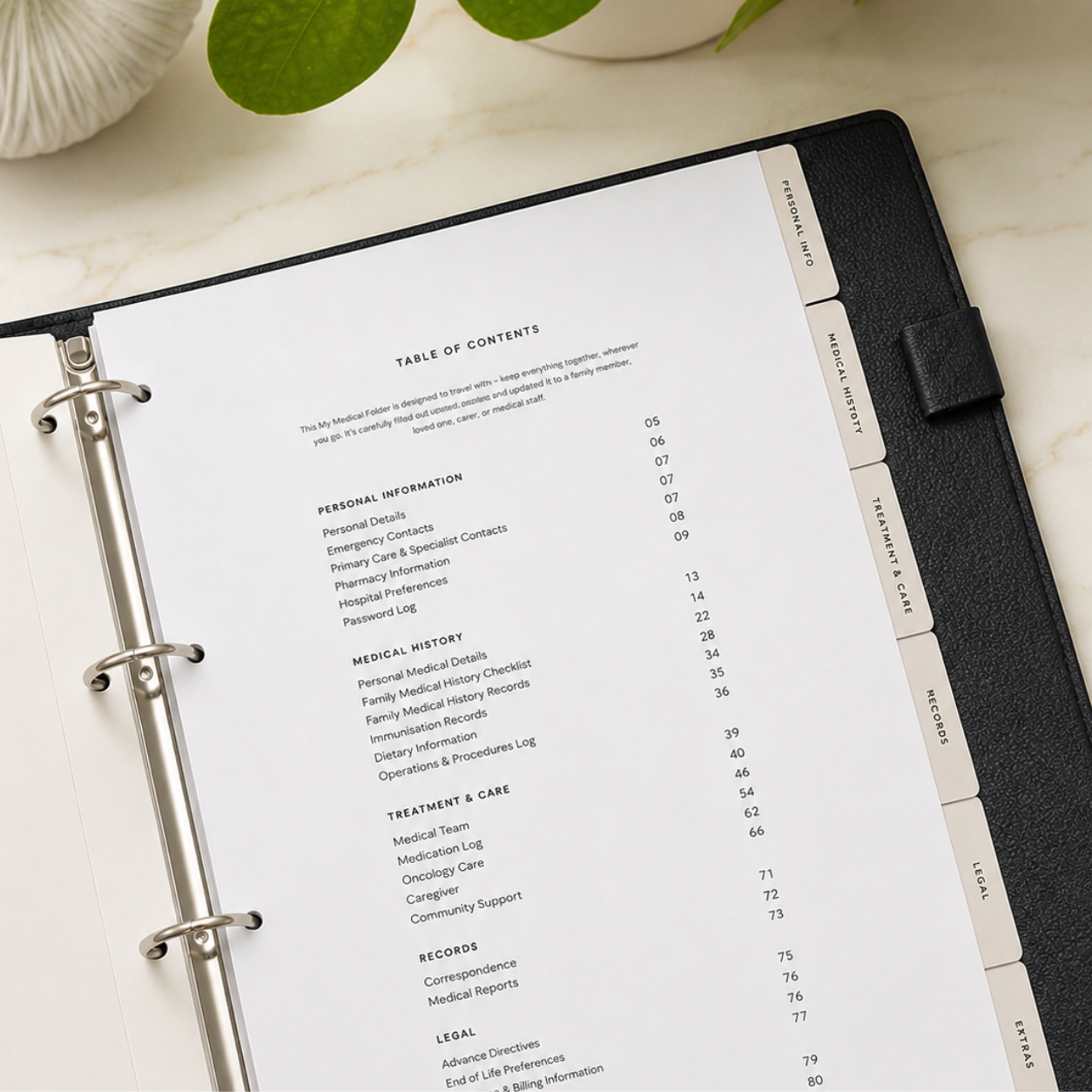 My Medical Folder open to the Table of Contents, showing organised health sections and labelled tabs on a cream marble surface with green foliage.